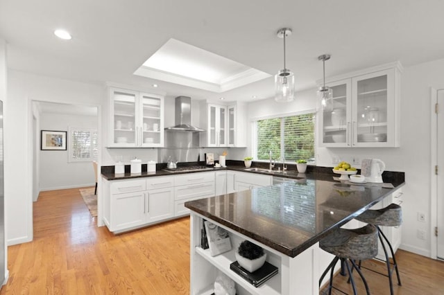 kitchen with sink, a breakfast bar area, white cabinets, hanging light fixtures, and wall chimney range hood