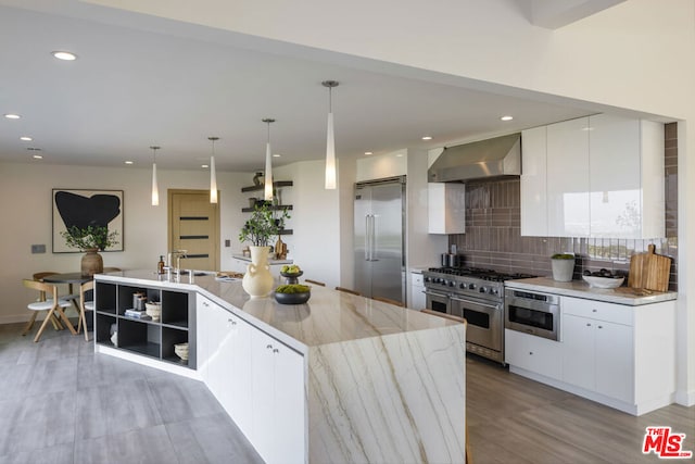 kitchen with white cabinetry, wall chimney exhaust hood, built in appliances, and decorative light fixtures