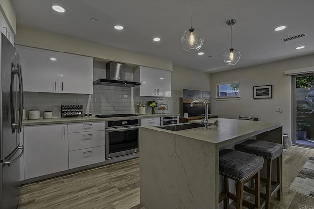 kitchen featuring appliances with stainless steel finishes, an island with sink, sink, white cabinets, and wall chimney exhaust hood