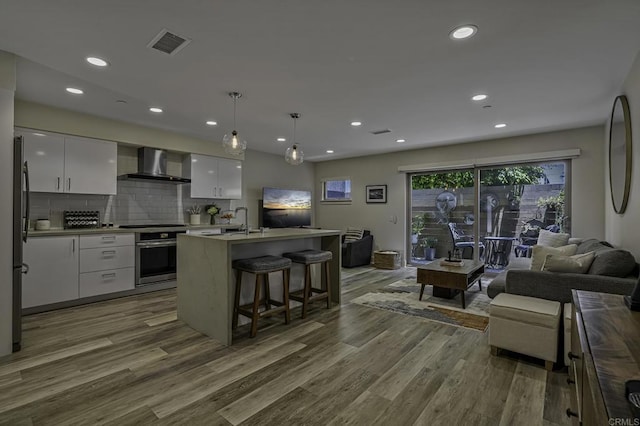 kitchen featuring decorative light fixtures, white cabinetry, oven, a kitchen island with sink, and wall chimney range hood