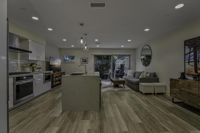 kitchen with sink, a kitchen island with sink, stainless steel appliances, white cabinets, and decorative light fixtures