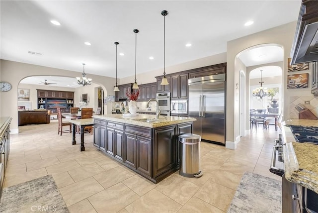 kitchen with built in appliances, pendant lighting, dark brown cabinetry, and an island with sink