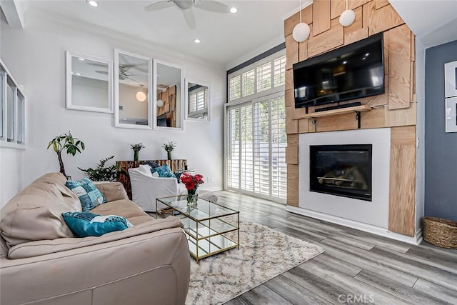 living room featuring wood-type flooring, ornamental molding, a large fireplace, and ceiling fan