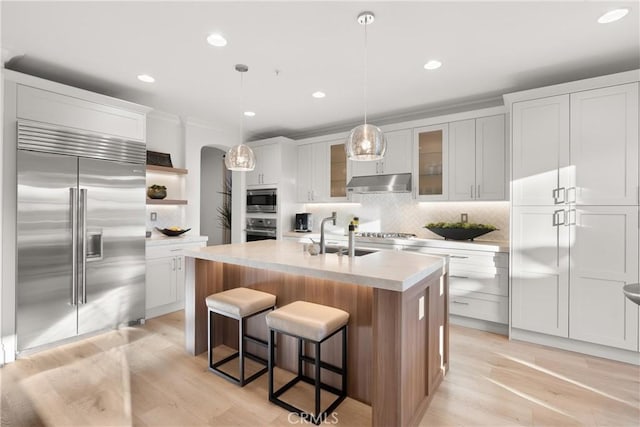 kitchen featuring white cabinetry, a kitchen island with sink, decorative light fixtures, built in appliances, and sink