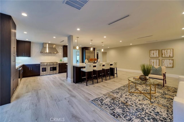 kitchen with wall chimney range hood, dark brown cabinets, stainless steel appliances, light hardwood / wood-style floors, and decorative backsplash