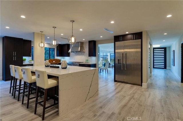 kitchen featuring backsplash, hanging light fixtures, high end appliances, light hardwood / wood-style floors, and wall chimney exhaust hood