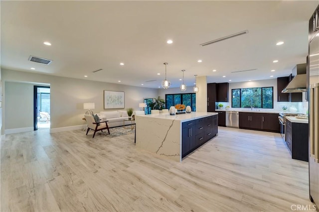 kitchen with appliances with stainless steel finishes, a kitchen island, pendant lighting, light hardwood / wood-style floors, and wall chimney range hood