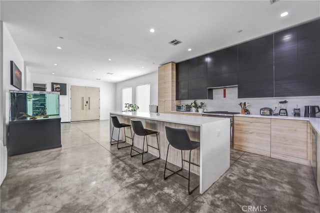 kitchen with a breakfast bar, light brown cabinetry, an island with sink, and decorative backsplash