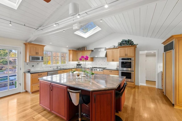 kitchen with lofted ceiling with skylight, a kitchen island, appliances with stainless steel finishes, light stone counters, and wall chimney range hood