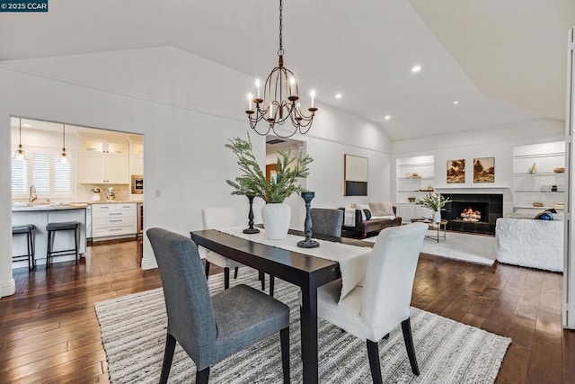 dining room with lofted ceiling, dark wood-type flooring, a tiled fireplace, built in shelves, and a chandelier