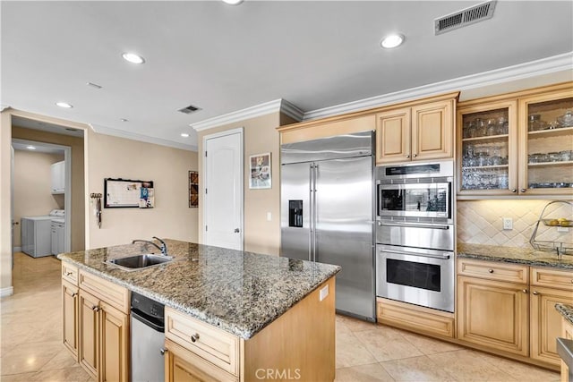 kitchen with appliances with stainless steel finishes, separate washer and dryer, sink, dark stone counters, and a kitchen island with sink