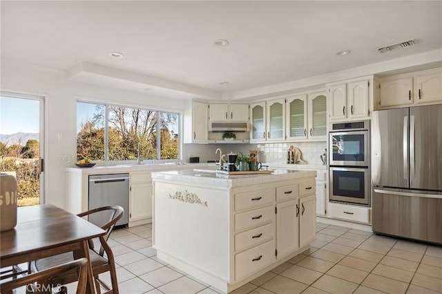 kitchen with white cabinetry, light tile patterned floors, an island with sink, stainless steel appliances, and decorative backsplash