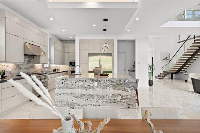 kitchen with tasteful backsplash, a center island with sink, modern cabinets, marble finish floor, and under cabinet range hood