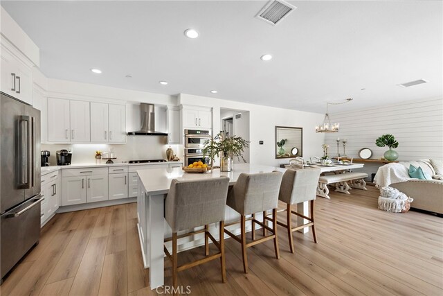 kitchen with wall chimney range hood, appliances with stainless steel finishes, white cabinetry, hanging light fixtures, and an island with sink