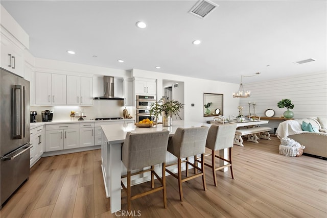 kitchen featuring wall chimney exhaust hood, decorative light fixtures, an island with sink, stainless steel appliances, and white cabinets