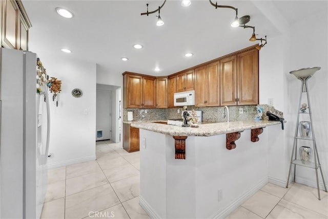 kitchen with white appliances, a breakfast bar area, backsplash, light stone counters, and kitchen peninsula