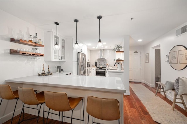 kitchen featuring wall chimney exhaust hood, kitchen peninsula, pendant lighting, stainless steel appliances, and white cabinets