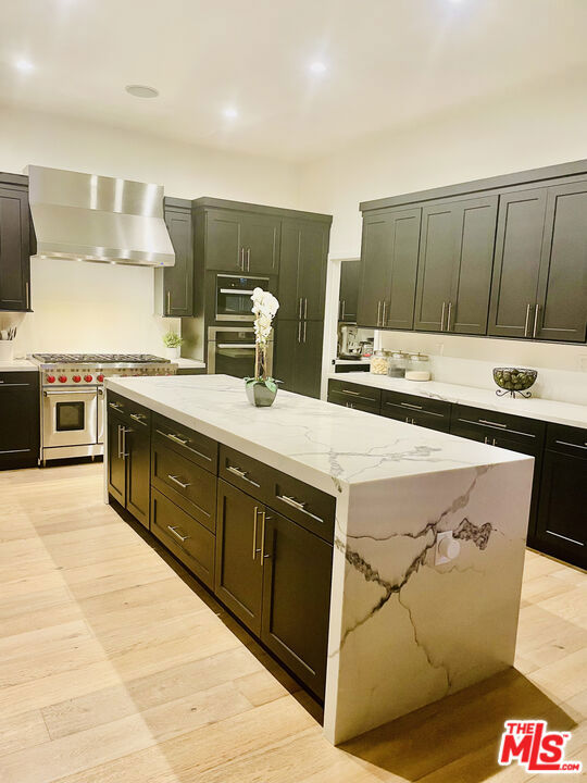 kitchen featuring wall chimney range hood, stainless steel appliances, light stone counters, light hardwood / wood-style floors, and a kitchen island