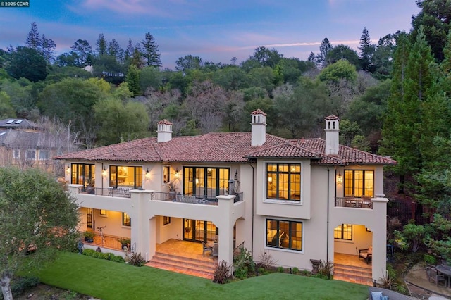 back house at dusk featuring a patio, a balcony, and a yard