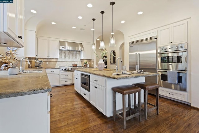 kitchen featuring white cabinetry, an island with sink, sink, built in appliances, and wall chimney exhaust hood