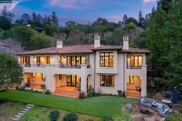 back house at dusk featuring a balcony, a yard, a patio area, and a fire pit