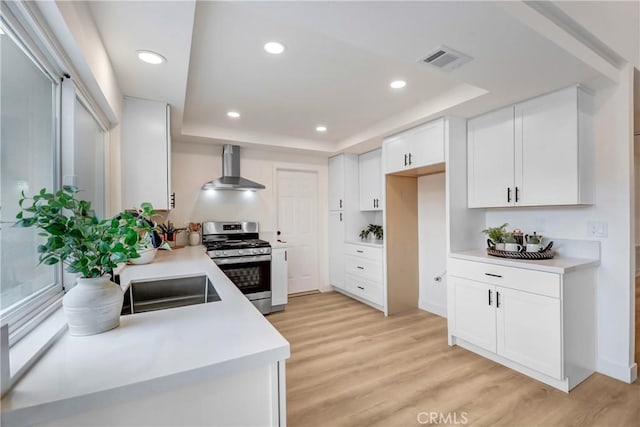 kitchen featuring wall chimney range hood, white cabinetry, gas range, a raised ceiling, and light wood-type flooring