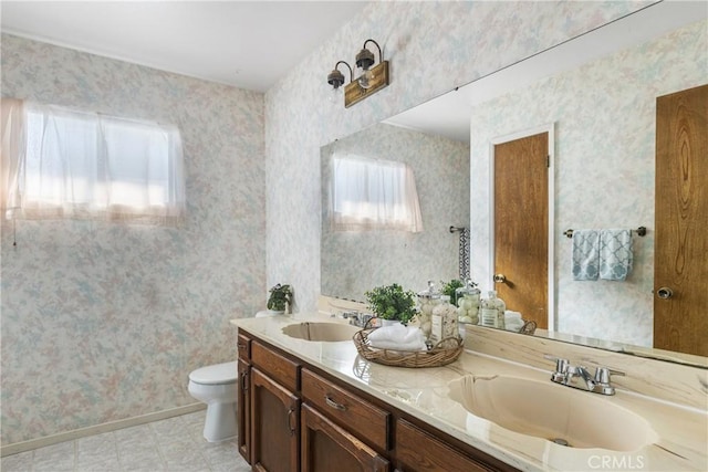 full bathroom with double vanity, a sink, toilet, and wallpapered walls