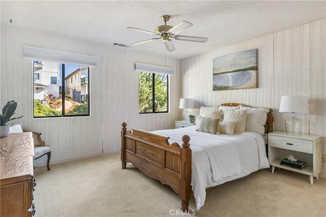 bedroom featuring light carpet, wallpapered walls, visible vents, a ceiling fan, and a textured ceiling