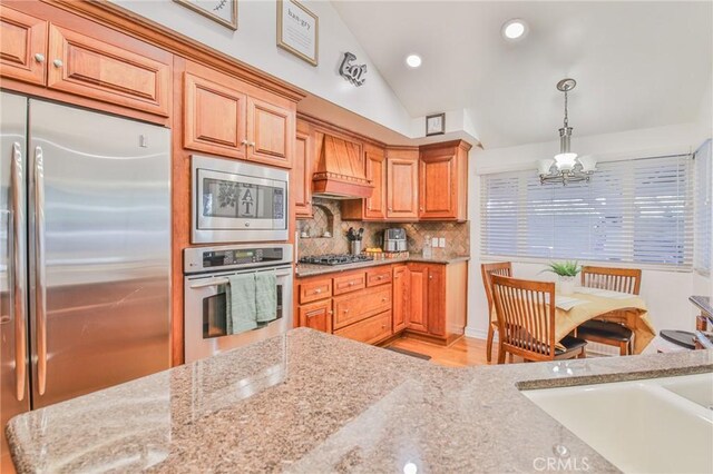 kitchen with vaulted ceiling, hanging light fixtures, stainless steel appliances, light stone counters, and custom exhaust hood