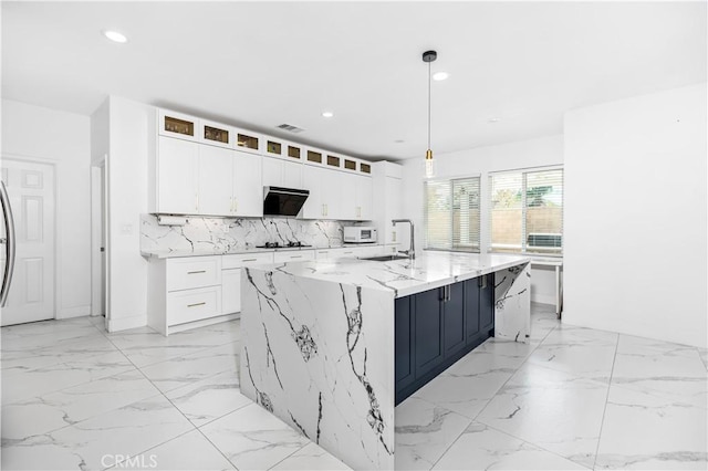 kitchen with light stone counters, white cabinetry, decorative light fixtures, a kitchen island with sink, and decorative backsplash