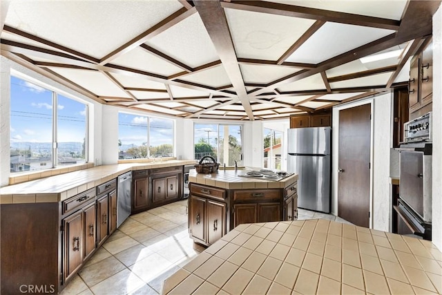 kitchen with dark brown cabinetry, coffered ceiling, a center island, appliances with stainless steel finishes, and tile counters