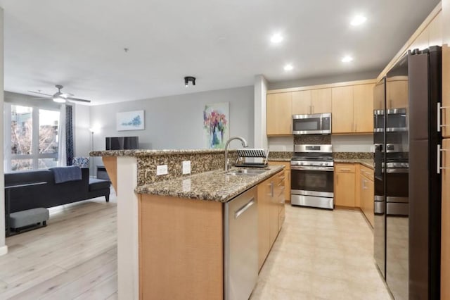 kitchen with sink, appliances with stainless steel finishes, dark stone countertops, an island with sink, and light brown cabinets