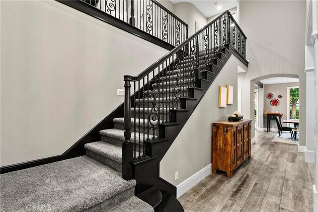 staircase featuring a towering ceiling, baseboards, arched walkways, and wood finished floors