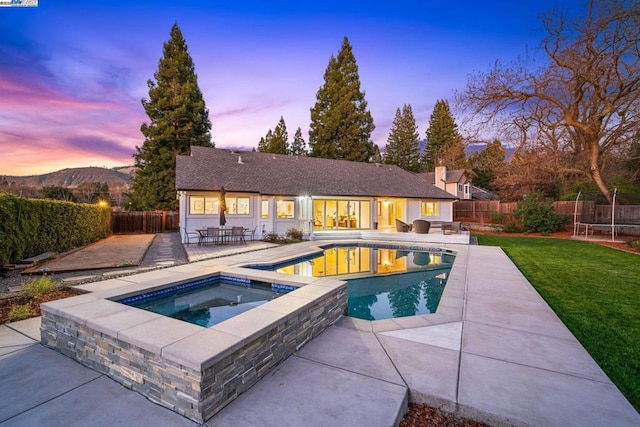 pool at dusk with a trampoline, a yard, a patio, a mountain view, and an in ground hot tub