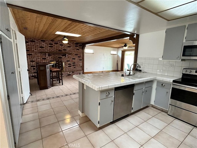 kitchen featuring sink, gray cabinetry, tile countertops, appliances with stainless steel finishes, and brick wall