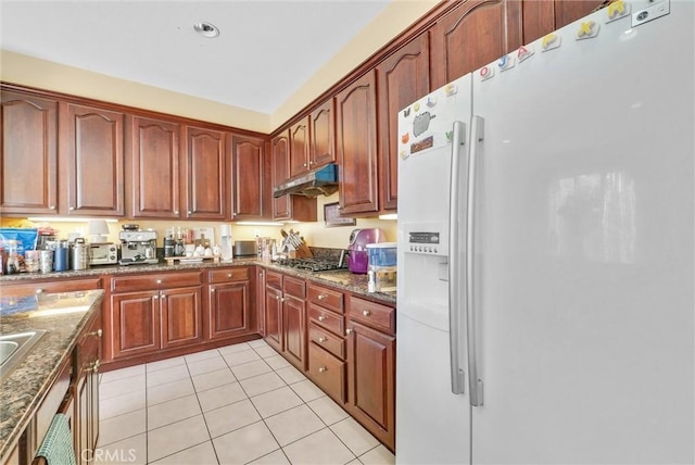 kitchen featuring stainless steel gas cooktop, white refrigerator with ice dispenser, light tile patterned floors, and dark stone countertops