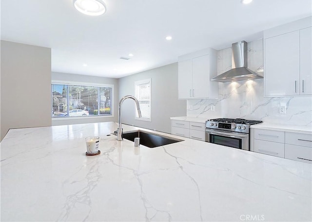 kitchen featuring wall chimney range hood, sink, stainless steel gas stove, light stone counters, and white cabinets