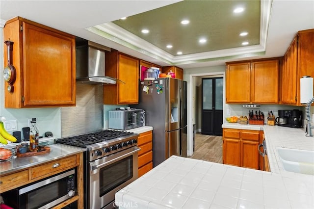 kitchen featuring a tray ceiling, tile countertops, stainless steel appliances, a sink, and wall chimney range hood