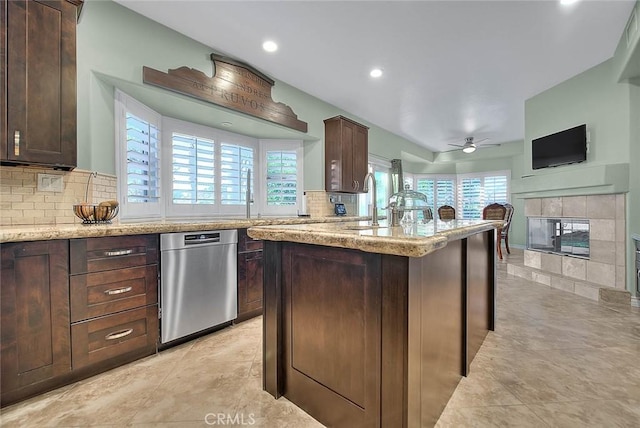 kitchen with dark brown cabinetry, an island with sink, light stone countertops, ceiling fan, and a tiled fireplace