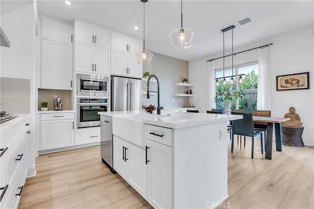 kitchen featuring appliances with stainless steel finishes, decorative light fixtures, white cabinetry, a center island with sink, and light wood-type flooring