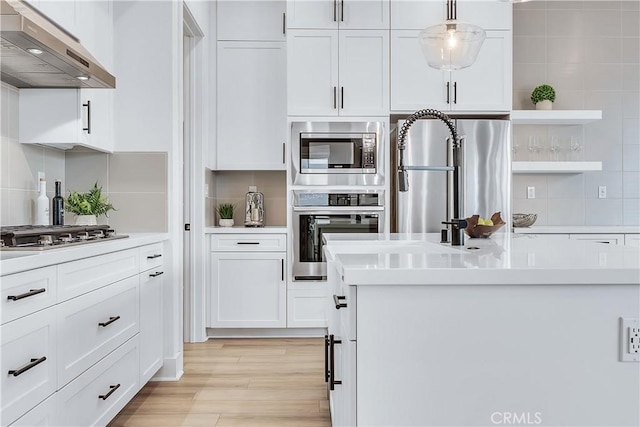 kitchen with appliances with stainless steel finishes, pendant lighting, white cabinetry, backsplash, and light hardwood / wood-style flooring