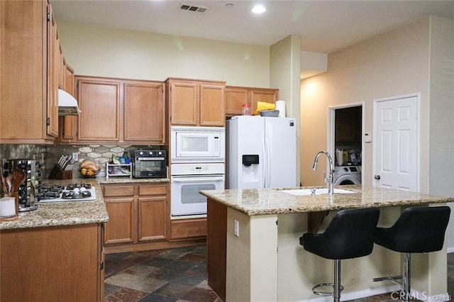 kitchen with sink, white appliances, light stone countertops, an island with sink, and decorative backsplash