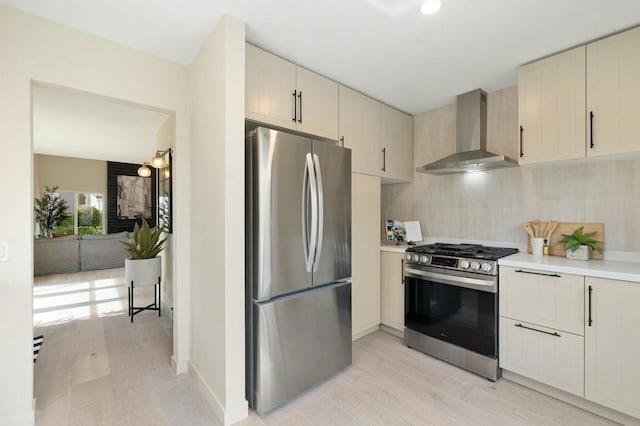 kitchen with appliances with stainless steel finishes, wall chimney range hood, cream cabinets, and light hardwood / wood-style floors