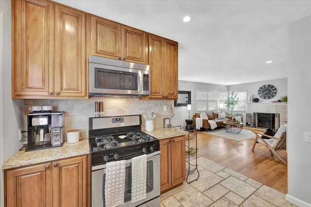 kitchen featuring a tile fireplace, backsplash, light hardwood / wood-style floors, stainless steel appliances, and light stone countertops