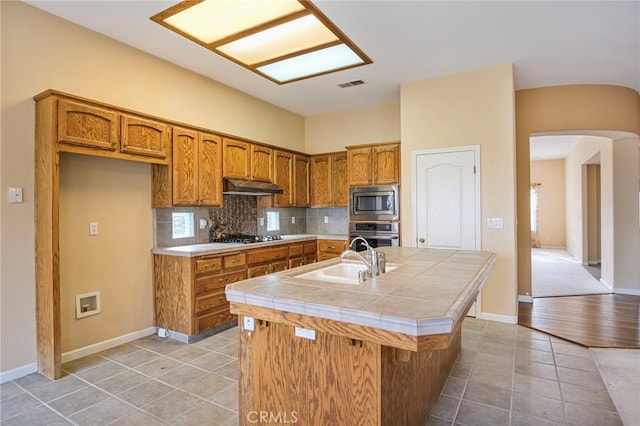 kitchen with tile countertops, sink, decorative backsplash, stainless steel appliances, and a center island with sink