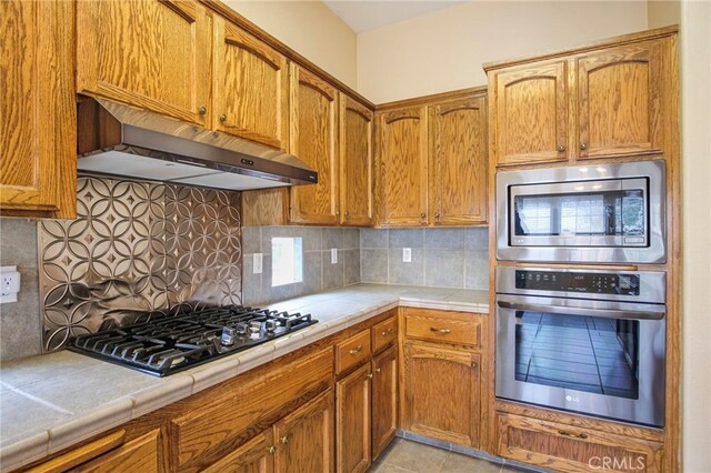 kitchen featuring stainless steel appliances, tile countertops, decorative backsplash, and exhaust hood