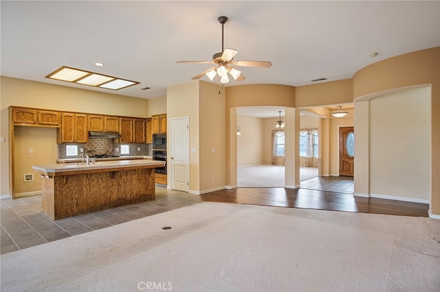 kitchen featuring ceiling fan, backsplash, a center island with sink, built in microwave, and oven