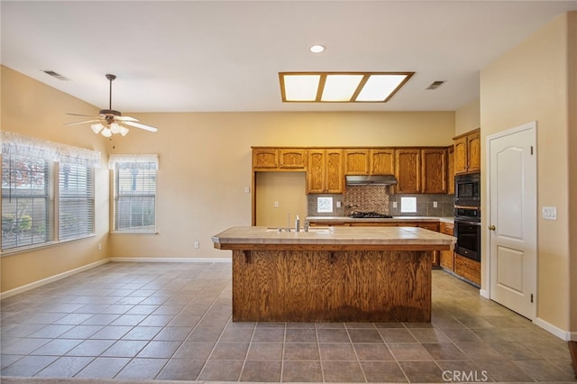 kitchen with black oven, built in microwave, an island with sink, tasteful backsplash, and stainless steel gas cooktop