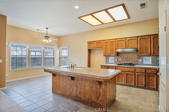 kitchen with sink, a center island with sink, tile counters, stainless steel gas stovetop, and backsplash