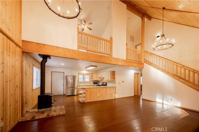 unfurnished living room with ceiling fan with notable chandelier, wood walls, lofted ceiling with beams, a wood stove, and dark hardwood / wood-style flooring